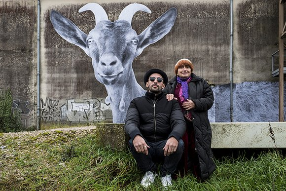A still from Visages, Villages: Varda and JR post in front of a large graffittied wall, upon which they've just posted a large mural of a goat with horns