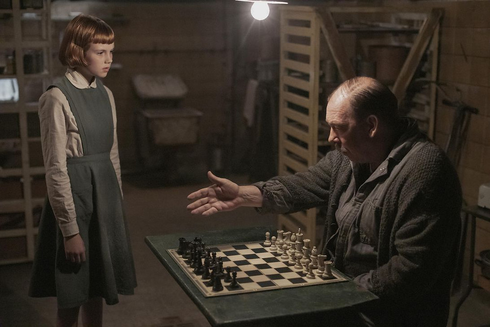 Elizabeth, as a child, stands before the caretaker and his chessboard as he gestures to the seat opposite.