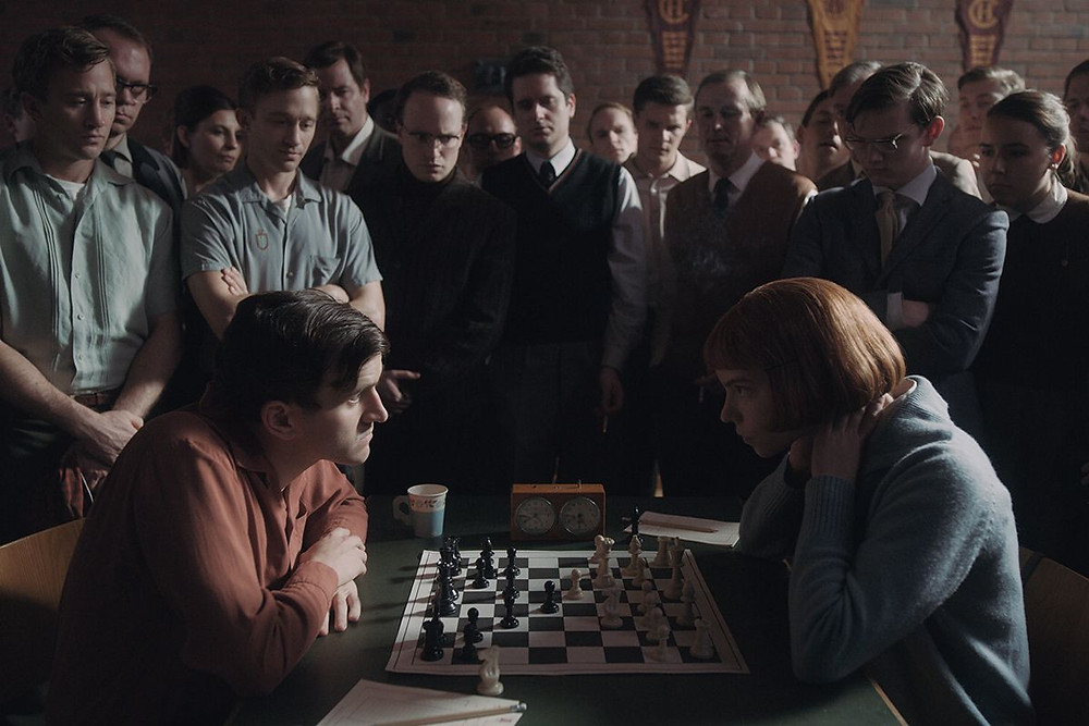 Elizabeth as a young woman plays a chess game against a champion, while a crowd of men watches.