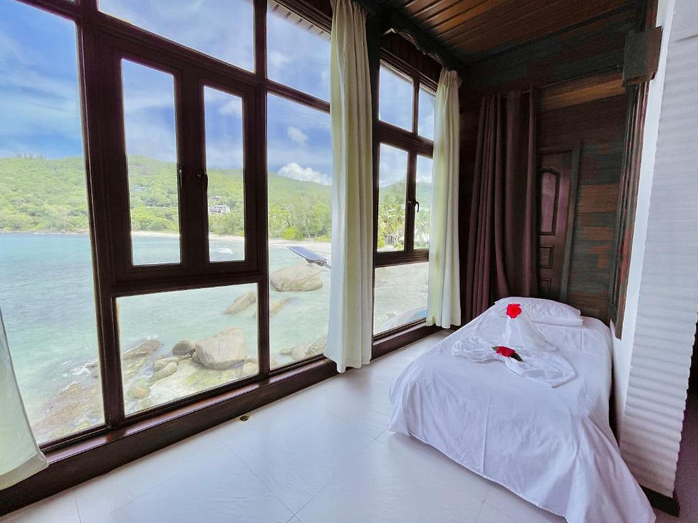 Cozy seaside spa room at Villa Chez Batista with a white massage bed, red flowers, and large windows overlooking turquoise water and green hills.