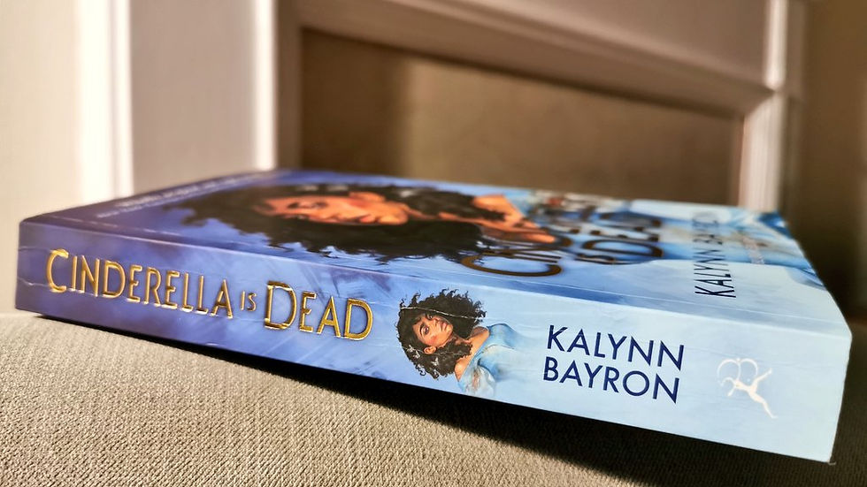 A photograph of the spine of the UK paperback of Cinderella is Dead by Kalynn Bayron. The book is balanced on the arm of a light grey armchair in front of a white and grey fireplace.
