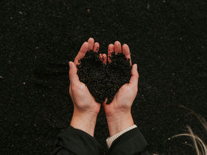Két kéz fekete homokot tart szív alakban, sötét háttér előtt. Meleg, földközeli hangulatot sugároz.