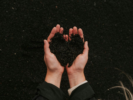 Két kéz fekete homokot tart szív alakban, sötét háttér előtt. Meleg, földközeli hangulatot sugároz.