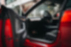 Red Tesla Model 3 interior with open door, showing black leather seats, wooden dashboard, and steering wheel. Suburban setting visible outside.