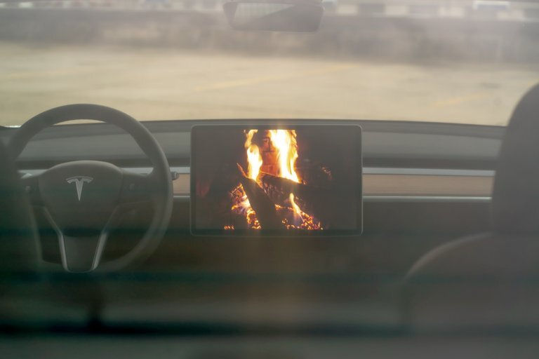 Innenansicht eines Tesla-Autos, Monitor zeigt virtuelles Kaminfeuer. Lenkrad sichtbar, Hintergrund unscharf. Stimmung warm und gemütlich.