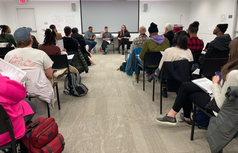 People seated in a circle in a modern room, engaging in a discussion. Notable: a red backpack, casual clothing, and a visible presentation screen.