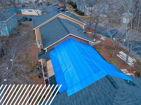 A house with a blue tarp covering part of the roof in a residential area with leafless trees. Supplies and a yard visible in the background.