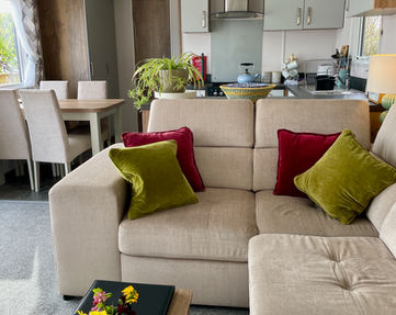Corner sofa facing a big TV next to the french doors opening onto the decking, seaview in the back. Behind the sofa is the kitchen and dining area.