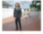 Groom in black suit stands on a patio, smiling, as bride in white dress approaches from behind. White fence and palm tree in background.