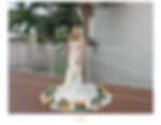 Bride in a white lace gown stands smiling, surrounded by colorful flowers. Palm trees and a railing in the background, creating a tropical vibe.