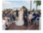 Bride and groom kiss at an outdoor wedding with guests seated on white chairs. The bride holds a bouquet; bridesmaids in green dresses nearby.