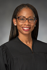 Black woman with braids smiling. She is wearing a black judges robe against a grey background.