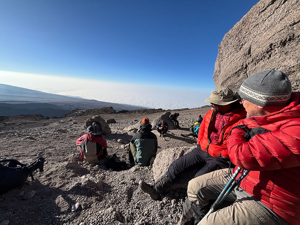 A group of climbers sitting on rocky terrain along the Rongai Route on Mount Kilimanjaro, relaxing and enjoying the spectacular panoramic views across the vast high-altitude landscape