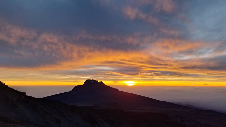 A breathtaking sunrise or sunset viewed from Mount Kilimanjaro along the Lemosho Route, with soft cotton-wool clouds below and warm colors illuminating the expansive high-altitude landscape.”