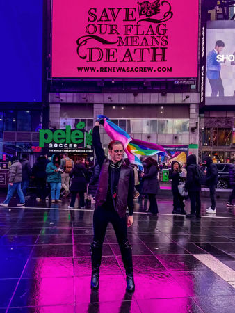 Photo by caribbeancrocs. OFMD Fan posing in Times Squar with the billboard while holding a rainbow fla & cosplaying a characted on the show.