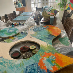 A colourful abstract painting on plyboard shape laying on a kitchen table.