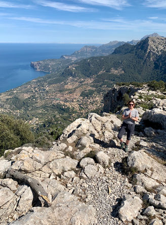 Fiona Marshall Yoga stands on mountain peak, overlooks scenic coastal view and town in Mallorca