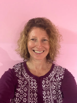 Smiling woman with curly hair poses in front of a pink wall in India, Fiona Marshall Yoga