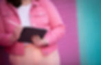 A close-up of a woman's hands holding a tablet and stylus, poised to write or draw. She wears a pink denim jacket and a smartwatch, with neatly manicured nails. The background features a colorful, pastel-brick wall, emphasizing a creative and modern aesthetic.