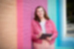 A woman with red hair leans against a vibrant, multicolored brick wall, holding a tablet and stylus. She is dressed in a pink denim jacket and a striped shirt, smiling warmly. The image captures a creative and professional atmosphere with bright, cheerful colors.
