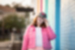 A woman with red hair smiles while adjusting a black baseball cap with a red heart emblem. She is wearing a pink denim jacket over a striped shirt and stands against a colorful, pastel-painted brick wall. The background is slightly blurred, with soft natural lighting.