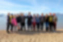 Group of people standing on the sandy shore of a lake with snowy mountains in the background