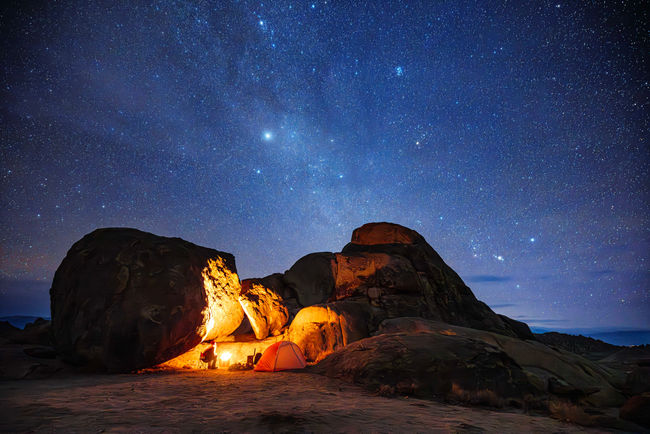 swirling stars in the night sky are visible over a vibrant rock formation and a warm campfire.
