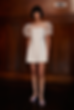 A person in a white dress with sheer sleeves stands against a wooden wall. They wear lilac shoes. The word "HONOR" is visible.