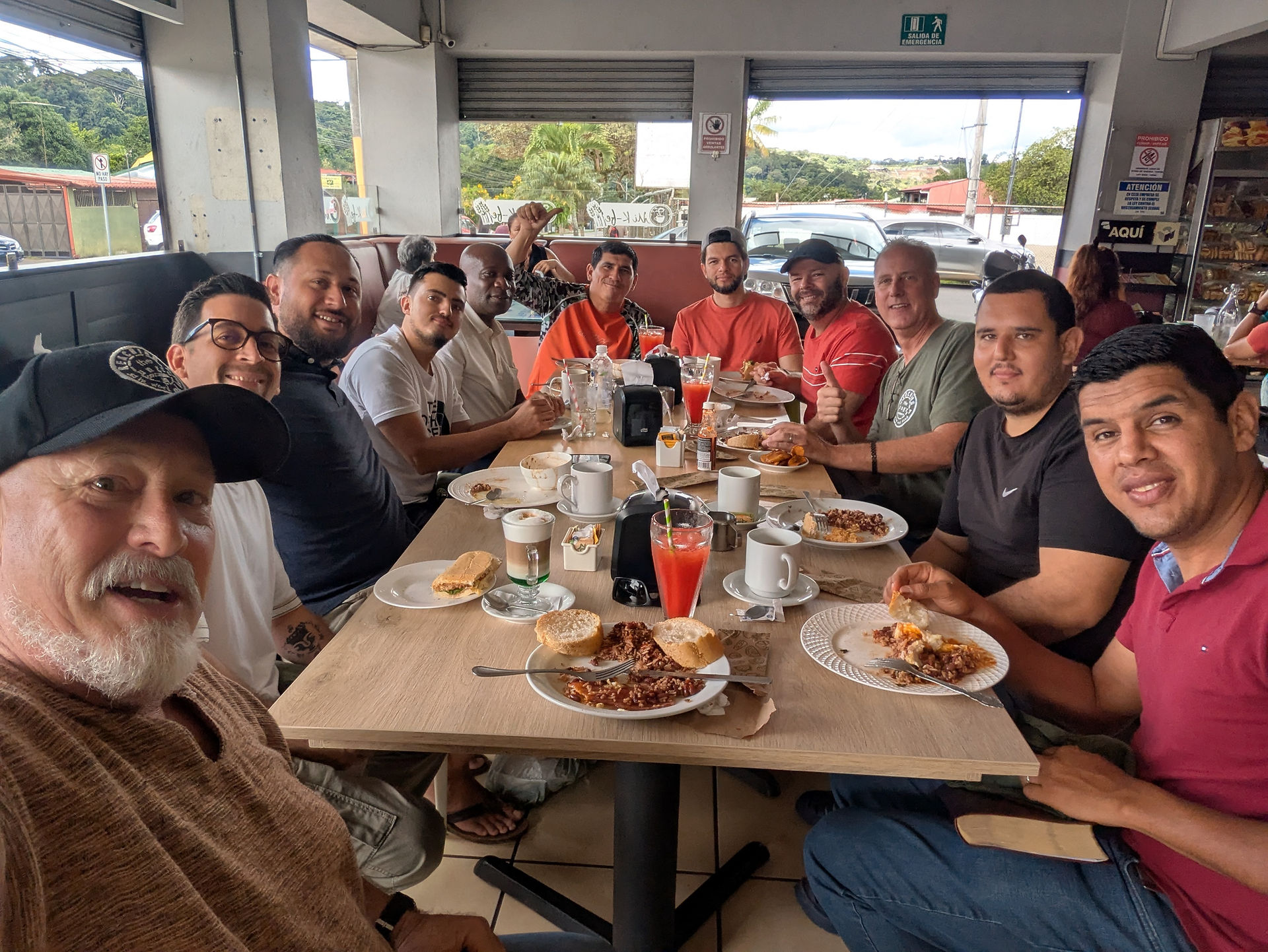 A group of men smiling and enjoying a meal. Plates of food and drinks on the table. Bright, casual setting with open windows.