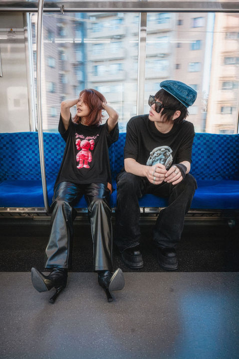 Two models sitting across from each other inside a Tokyo subway carriage wearing oversized graphic streetwear t-shirts, captured through a moving train window with city buildings blurred outside, photographed by Tokyo photographer Deniz Demir.