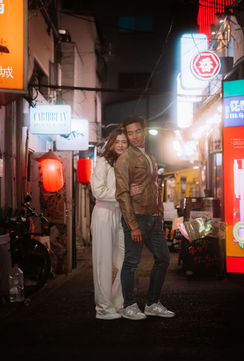 A couple stands together in a quiet Tokyo alley, posing for a photo. The woman wears a white sweater and the man is dressed casually. The alley is lit by soft streetlights, with vibrant neon signs visible in the background.