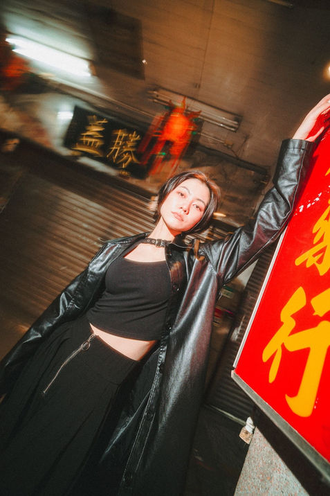 A woman in a black leather coat rests her arm against a bright red sign in a narrow market street at night, neon reflections blurred behind her. She meets the camera with a steady expression, illuminated by warm storefront light. Shot in Taipei