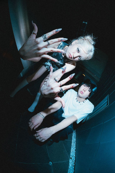 Two models sit close together in a dim urban corridor, extending their hands toward the lens to showcase silver rings and long, sculptural nails. The wide-angle perspective and flash lighting amplify a gritty, futuristic Japanese fashion atmosphere. Shot by Tokyo photographer Deniz Demir for Fragile Base and Yokogao Mag.