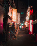 Two friends walk down a brightly lit alley in Tokyo at night, surrounded by colorful neon signs and glowing lights. One friend wears a leather jacket and the other smiles as they share a moment together.