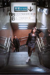 A woman dressed in a black jacket, skirt, and boots walks up the stairs at Shinjuku Station in Tokyo. The bright sign above indicates the direction to Shinjuku Terminal, while another person follows her up the stairs. The scene is illuminated with soft lighting, creating a dynamic, urban atmosphere.