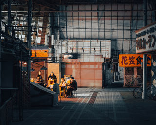  A group of people sits at a restaurant in Tokyo, illuminated by warm lights in a quiet alley. The empty street surrounding the restaurant adds to the cozy ambiance of the place, enhancing the nighttime vibe. NFT Photography by Tokyo Photographer Deniz Demir. Tokyo, Japan.