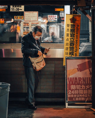  A man in a gray coat checks his phone while standing near a security booth in a Tokyo alley, surrounded by signs warning about surveillance. The city's bright signage and neon lights provide an urban, cinematic feel to the photo. NFT Photography by Tokyo Photographer Deniz Demir. Tokyo, Japan