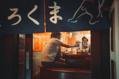 People sit at a narrow wooden counter inside a small Tokyo izakaya, seen through a partially open noren curtain with Japanese lettering. Warm interior lighting contrasts with the dark street outside, emphasizing intimate night dining culture. Night street photograph by Tokyo photographer Deniz Demir.