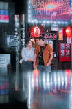 A couple smiles at each other while holding hands in front of a neon-lit restaurant in Tokyo. The scene is illuminated by glowing red lanterns and vibrant neon signs, creating a romantic and lively atmosphere in the bustling city.