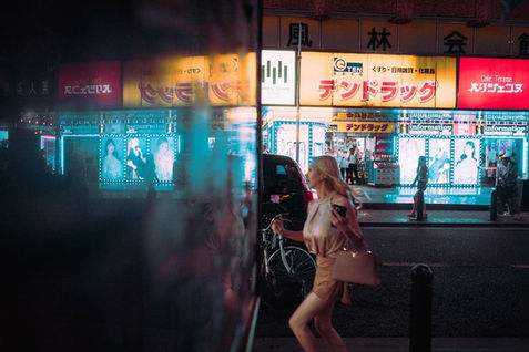 A woman walks past brightly lit storefronts and illuminated advertisements in a Tokyo entertainment district at night. Reflections in glass partially obscure the scene, blending interior light with the street outside. Night street photography by Tokyo photographer Deniz Demir.