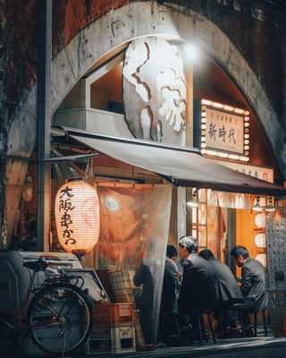 A dimly lit alley in Tokyo with people seated at a restaurant beneath a large sign and glowing lanterns. The scene is captured in a warm, inviting glow from the lanterns and restaurant lights, with people in business attire enjoying a meal.NFT Photography by Tokyo Photographer Deniz Demir. Tokyo, Japan.