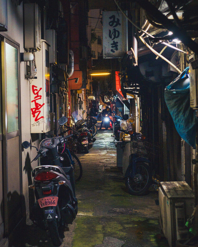 A narrow alleyway at night lined with parked motorbikes, glowing signs, and small shops, leading to a warmly lit area in the distance.