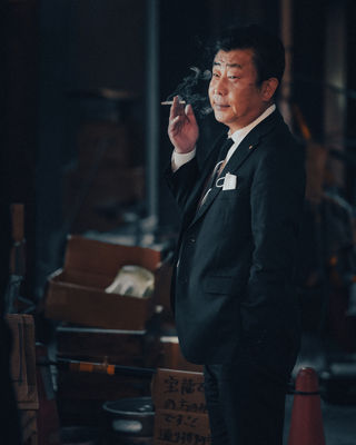 A businessman in a black suit and white shirt stands outside, exhaling cigarette smoke. The background features blurred elements of Tokyo's street life, creating a moody, atmospheric feel. He exudes a calm, reflective demeanor.