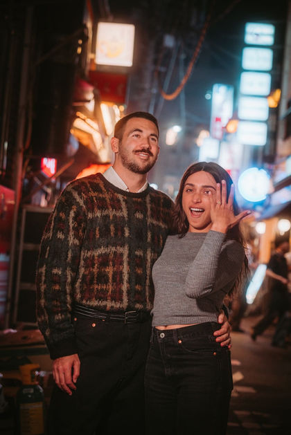 A newly-engaged couple smile in a narrow lantern-lit alley at night. The woman holds her hand forward, proudly showing her engagement ring, while her partner wraps an arm around her waist. Blurred signs and warm lights give a cozy Tokyo nightlife atmosphere. Shot by Tokyo portrait and couples photographer Deniz Demir.