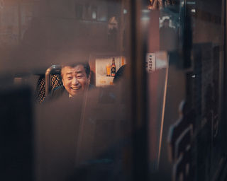A man can be seen smiling through the window of a Tokyo restaurant, his reflection captured on the glass. The scene is intimate, with the soft glow of the restaurant light contrasting with the outdoor environment. NFT Photography by Tokyo Photographer Deniz Demir. Tokyo, Japan.