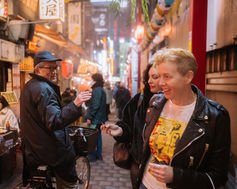 A man on a bicycle talks to a woman in a busy alley in Tokyo, where small food stalls and shops line the streets. The area is illuminated by hanging lanterns and streetlights, adding to the lively ambiance.