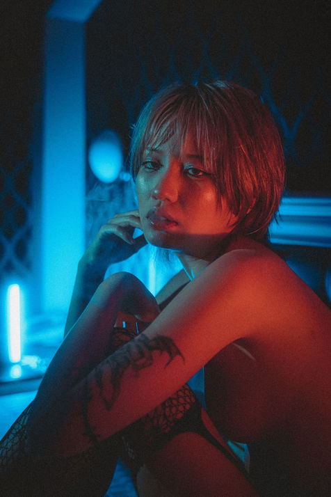 Close portrait of a model seated on a bed under contrasting blue and red lighting. Her short blonde hair and tattoos catch subtle highlights as she rests a hand near her face, the moody color palette lending a cinematic feel.