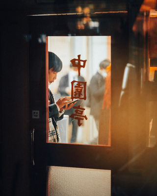 A young man stands inside a restaurant, holding a smartphone and engrossed in his device. The photo captures a partial view of the bustling interior through the restaurant's glass door, with the Japanese characters on the door adding a cultural touch to the urban scene.