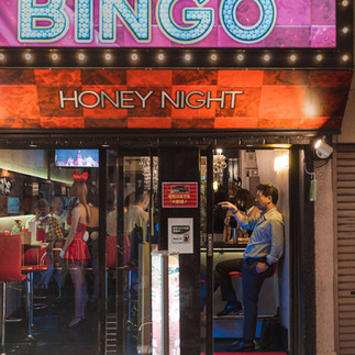 A bar entrance with neon signs reading "BINGO" and "HONEY NIGHT." Inside, people sit at the counter while a woman in red stands near the bar.