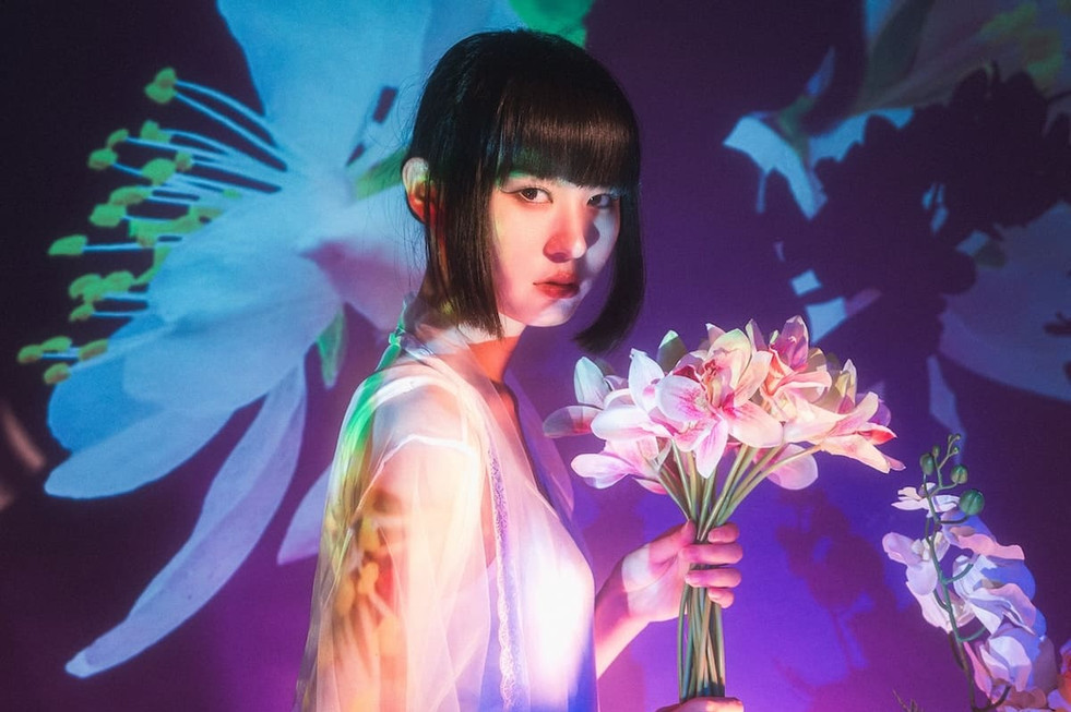 A woman with a sleek black bob holds a bouquet of pink and white orchids while standing in front of a projection of oversized white flowers. Her translucent lace robe glows under vivid hues of green, pink, and purple lighting, creating a surreal and ethereal mood.
